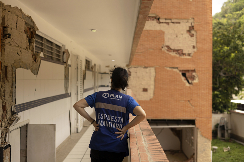 A woman standing on a balcony of a building damaged by earthquakes in Venezuela faces away from the camera. She is wearing a Plan-branded blue t-shirt that reads, “espuesta humanitarian,” which translates to “humanitarian response,” on the back.