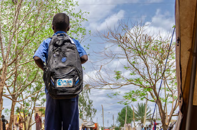 A young boy stands outside a tent in a displacement camp in Sudan, wearing a Plan-branded backpack. He is one of 1,000 children in Sudan who have received school kits from Plan. 
