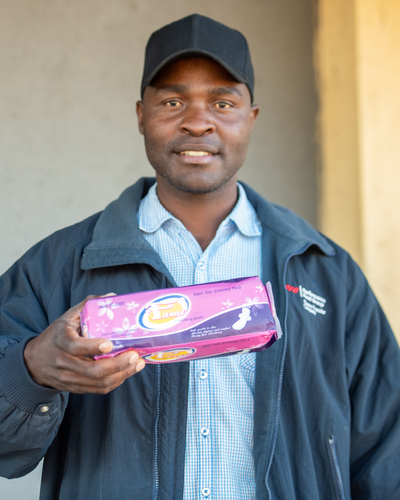 A man in a black baseball cap and blue jacket poses with a box of menstrual pads in his hand.