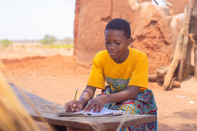 A girl in a yellow shirt and dress, sitting at desk writing in a notebook outside