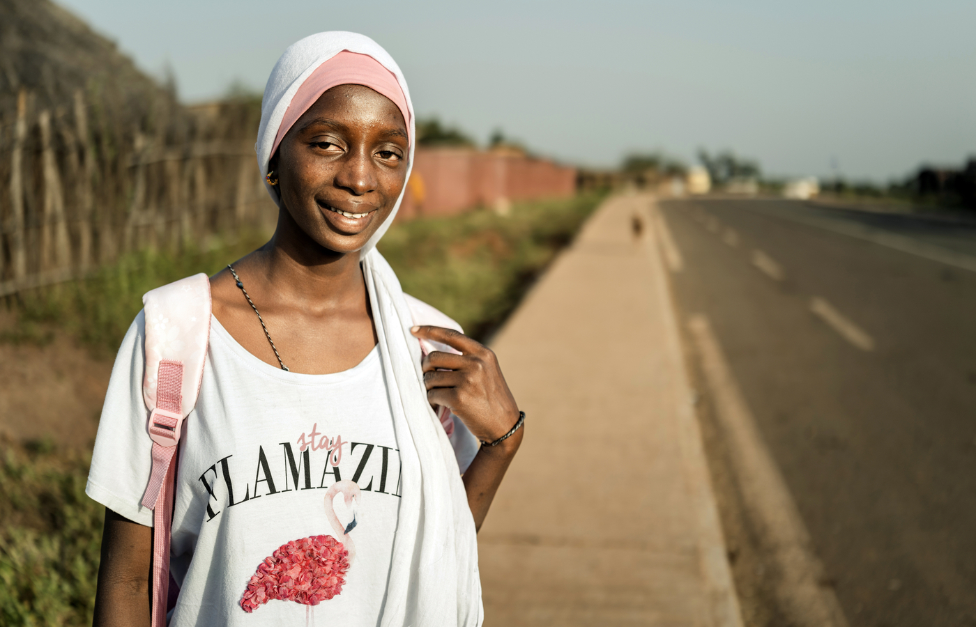 A smiling teenage girl wearing a white and pink head scarf and a white t-shirt stands on the sidewalk beside a road in rural Senegal.