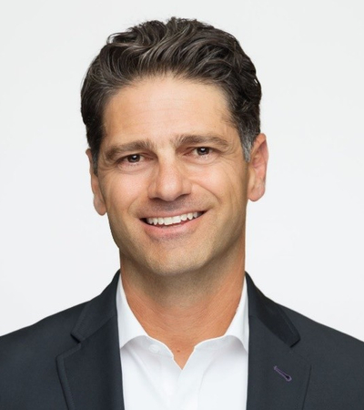 A professional headshot of a man wearing a dark blazer over a white collared shirt. The background is plain light gray.