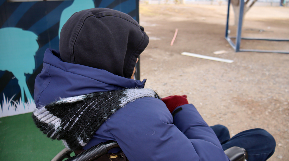 A teenage boy wearing a thick blue jacket and hat is photographed from behind. He is outdoors, sitting in a wheelchair.