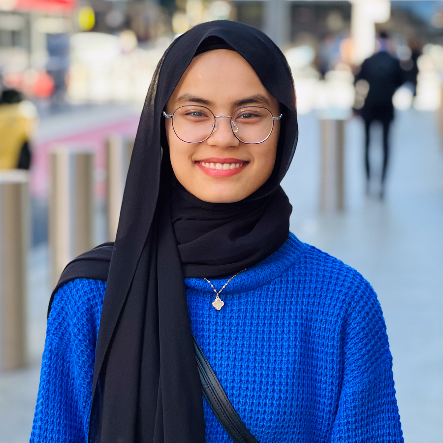 A young woman wearing a blue sweater and a black headscarf smiles at the camera. She is wearing glasses and is standing outdoors. The background is blurry.