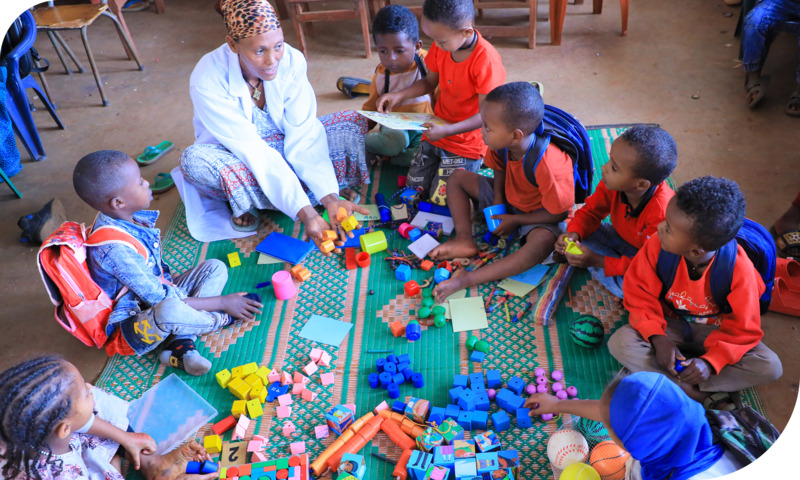 A group of young children and their teacher sit in a circle on the floor; blocks, balls and other toys are spread out in front of them.