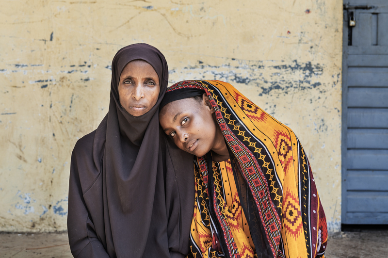 An older woman in a brown hijab looks straight into the camera as a younger woman in a yellow, black, and red headscarf leans on her shoulder, looking into the camera.