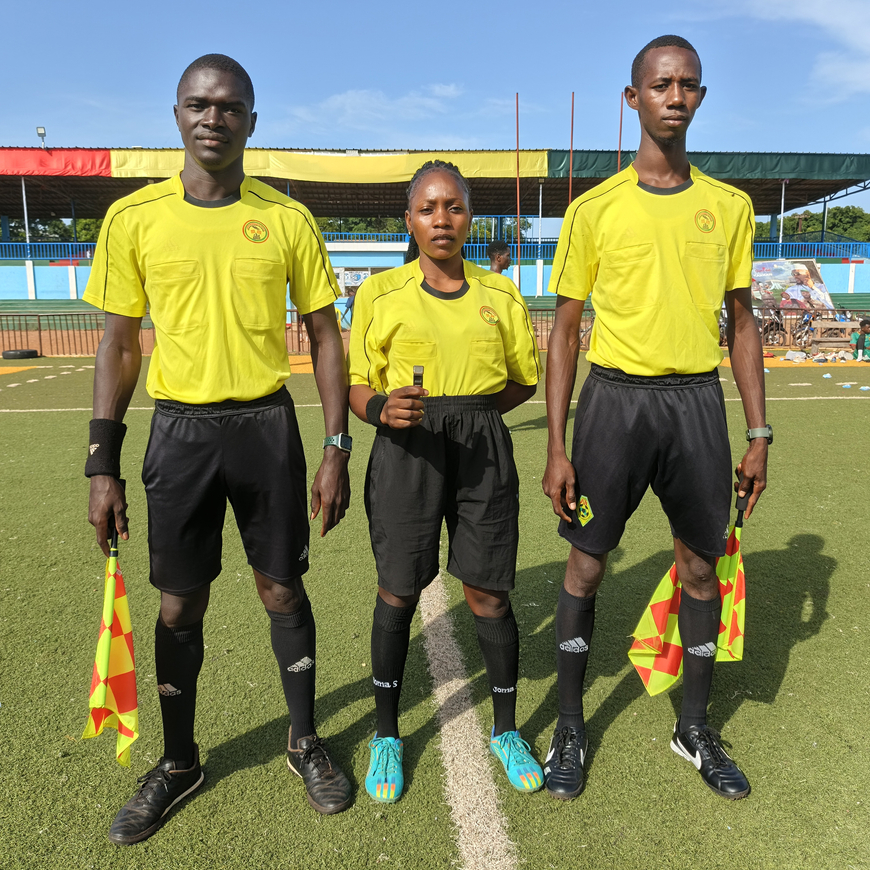 Three referees stand side by side on a football field. A woman wearing a yellow t-shirt and black shorts stands between two men wearing the same uniform. All three are facing the camera, with the field visible behind them.
