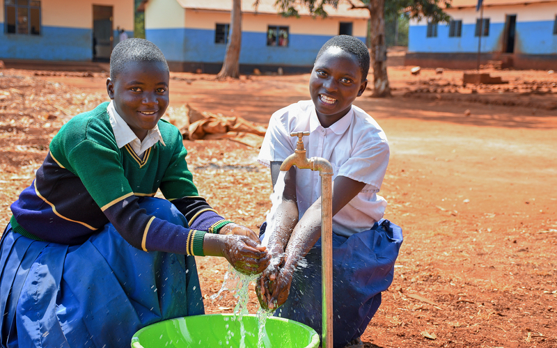 Two schoolgirls washing their hands at an outdoor spigot, representing how access to clean water supports girls’ health and education.
