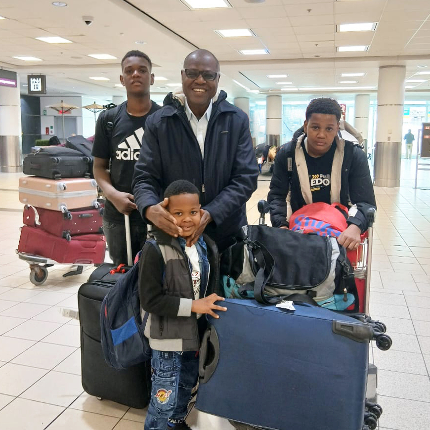 A man and three boys stand together in an airport with trolleys piled with suitcases.