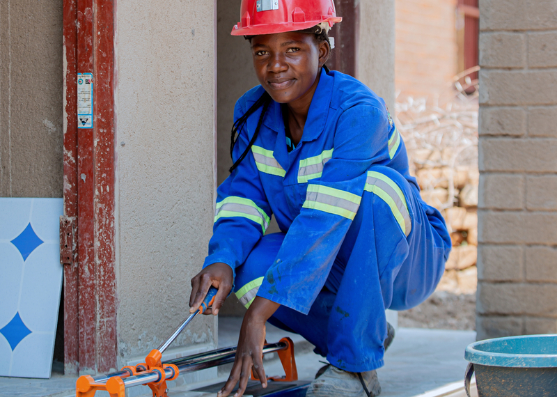 A young woman in a blue construction outfit, cutting a metal pipe outside a home she is building.