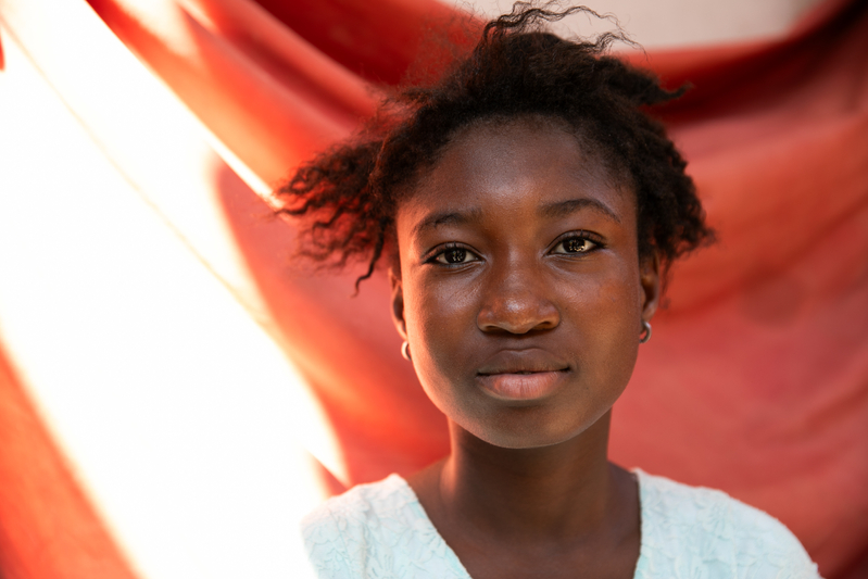 An adolescent girl looks directly at the camera. She stands against a hanging orange cloth that is partially bathed in sunlight.