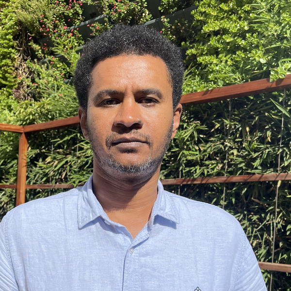 A man wearing a light blue shirt standing outside in the sunlight surrounded by green trees.