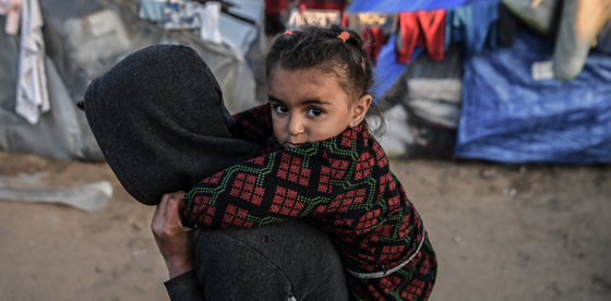 A photo of a young child looking directly into the camera as they’re being carried on the shoulders of another youth in Rafah.