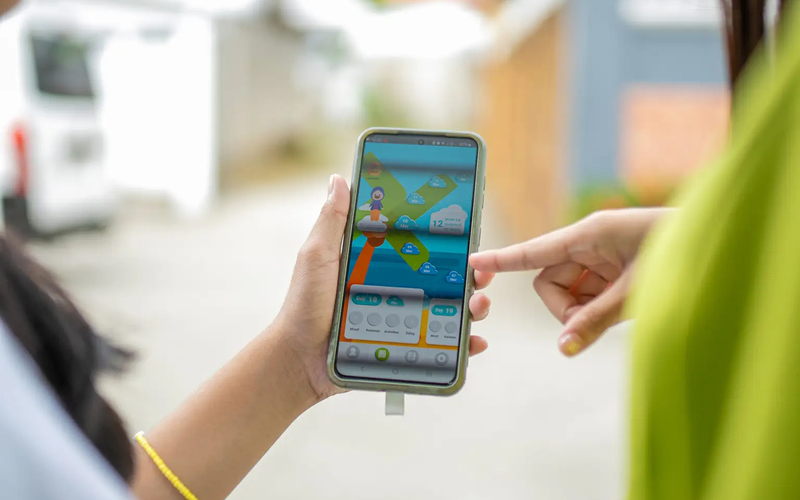 A zoomed- in photo of the hands of two people, one holding a smartphone and the other pointing to the screen which displaysing contents of an app. graphics.
