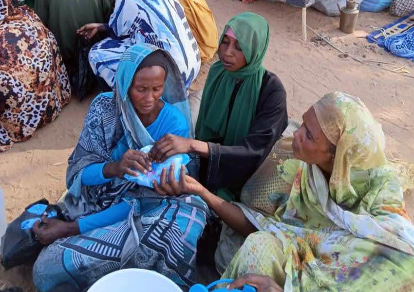 A group of three women surrounded by makeshift tents in a sandy area.
