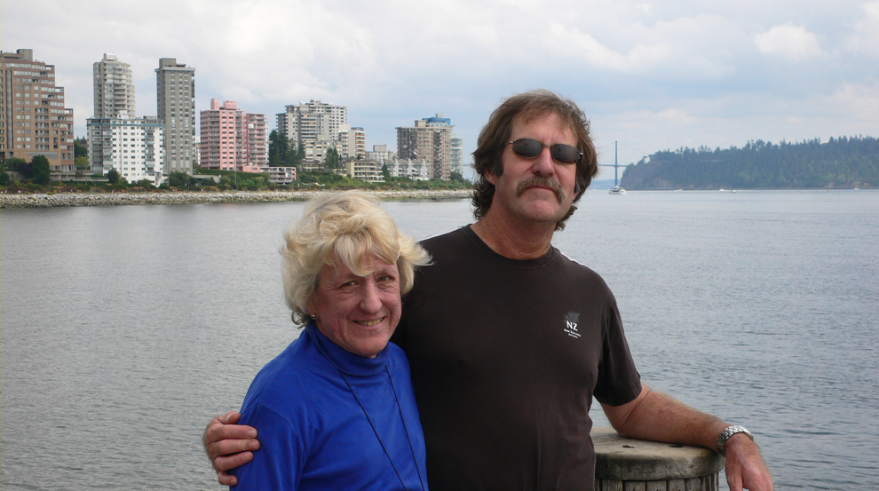A woman in a blue turtleneck stands next to a man in a black t-shirt with sunglasses in front of a waterfront with buildings and a bridge in the background.