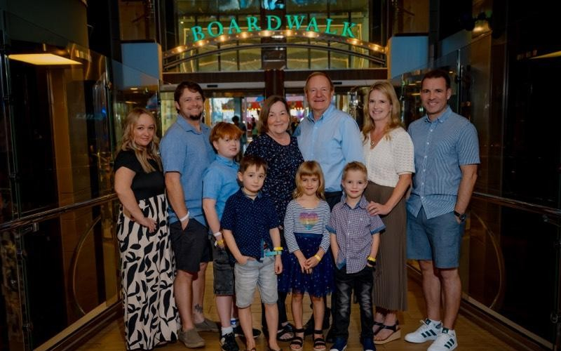 A family photo of 10 people, including four children, posing in front of a neon green sign that reads “boardwalk” inside a cruise ship.