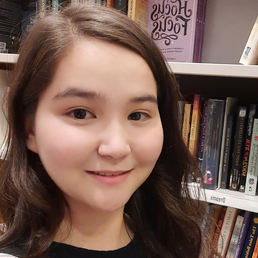 A young woman stands in front of a shelf of books. She has a white strap on one shoulder and has medium brown hair.