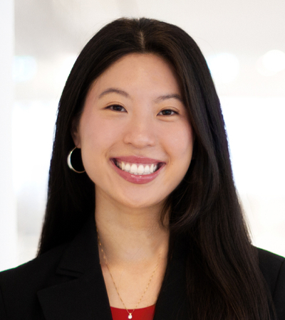 A portrait of a young woman wearing a black blazer over a red shirt. She is standing indoors against a softly blurred office background, facing the camera.