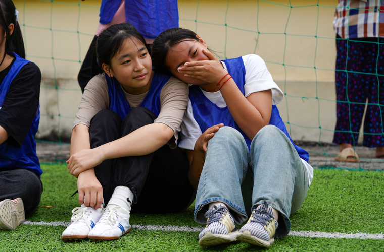  Two young women sit on a grassy field, one comforting the other, who appears emotional. They wear sports vests and casual clothing.