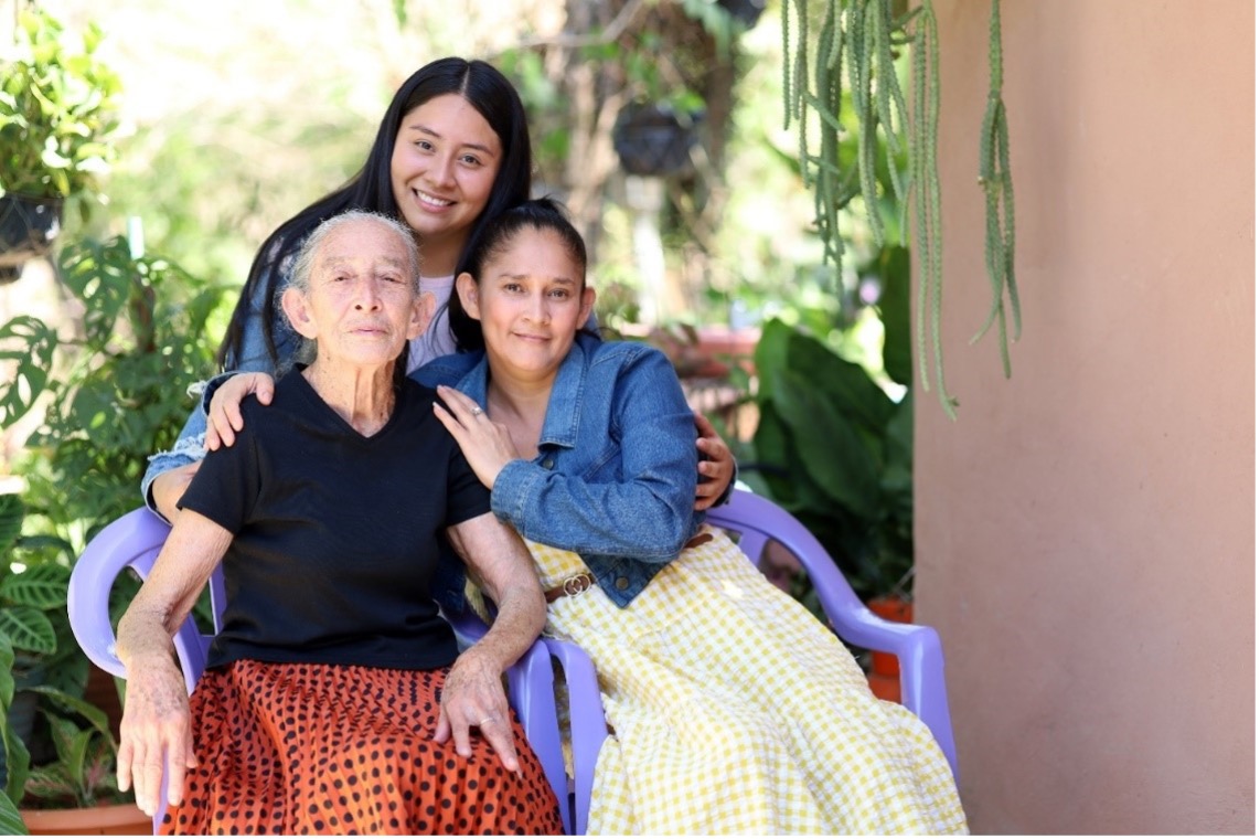 Two women sitting on purple chairs while a girl hugs them around the shoulders from behind