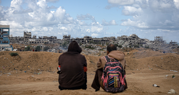 Two people sit on a sandy dune in the Al-Sudaniya beach area, overlooking a devastated cityscape, with collapsed buildings and rubble stretching toward the horizon under a partly cloudy sky.