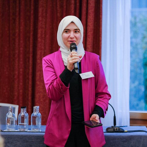 A teenage girl wearing a pink blazer and white hijab speaking with a microphone in her hand.