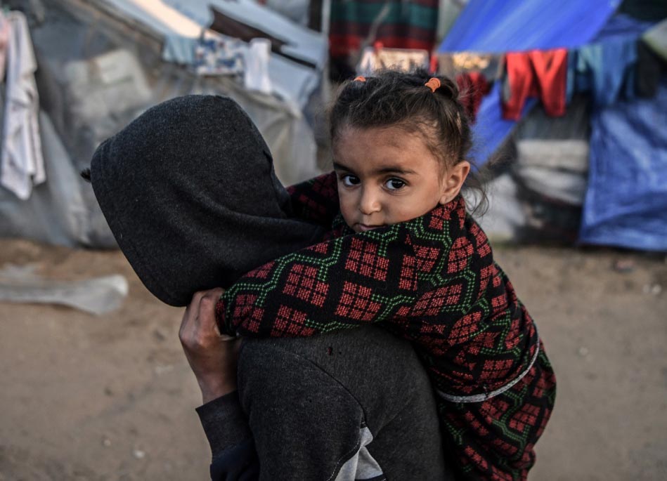 A photo of a young child looking directly into camera as they’re being carried on the shoulders of another youth in Rafah.
