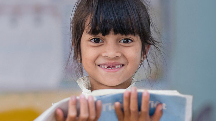 AA smiling woman stands in the centre of the picture, holding a small plastic bag that contains a seedling. She is surrounded by other women who are bent over tending to other seedlings.