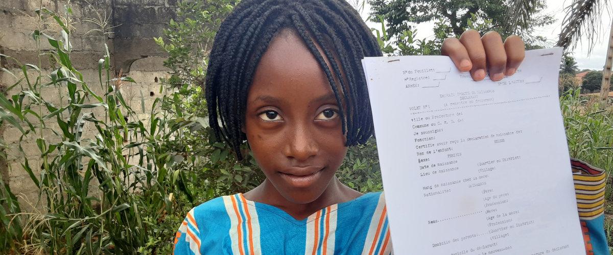 A young girl in a colorful dress holds a document, standing outdoors with greenery in the background.