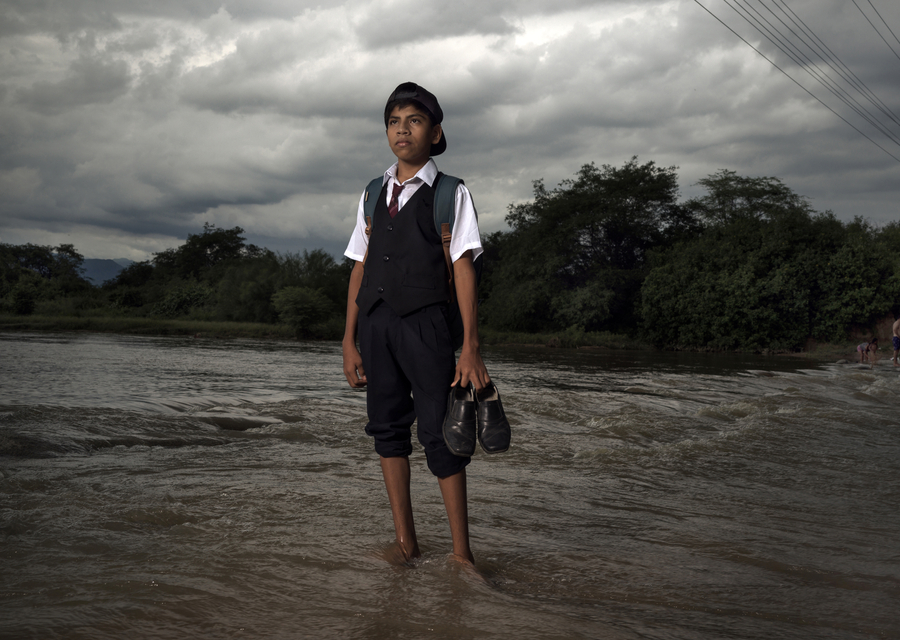 A boy in a school uniform stands barefoot in a river, holding his shoes, highlighting the dangerous journey to school during Peru’s rainy season.
