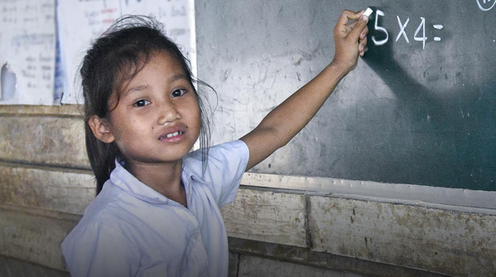A smiling teenage girl wearing a white and pink head scarf and a white t-shirt stands on the sidewalk beside a road in rural Senegal.