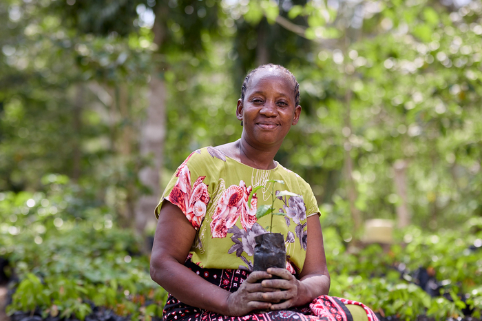 A woman looks into the camera smiling, while she is sitting down holding a small seedling in a black plastic bag.