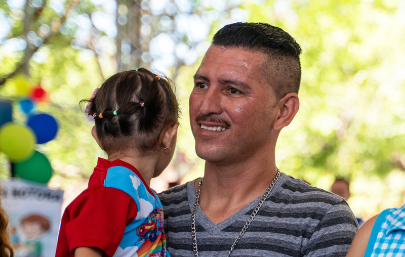 A man kneels down next to his young daughter, who is sitting in a swing at a playground.