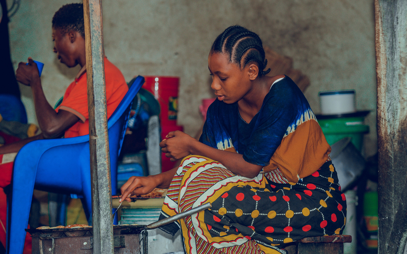 A girl in traditional clothing sits in a cafe with a woman in the  background wearing red sitting on a blue chair.