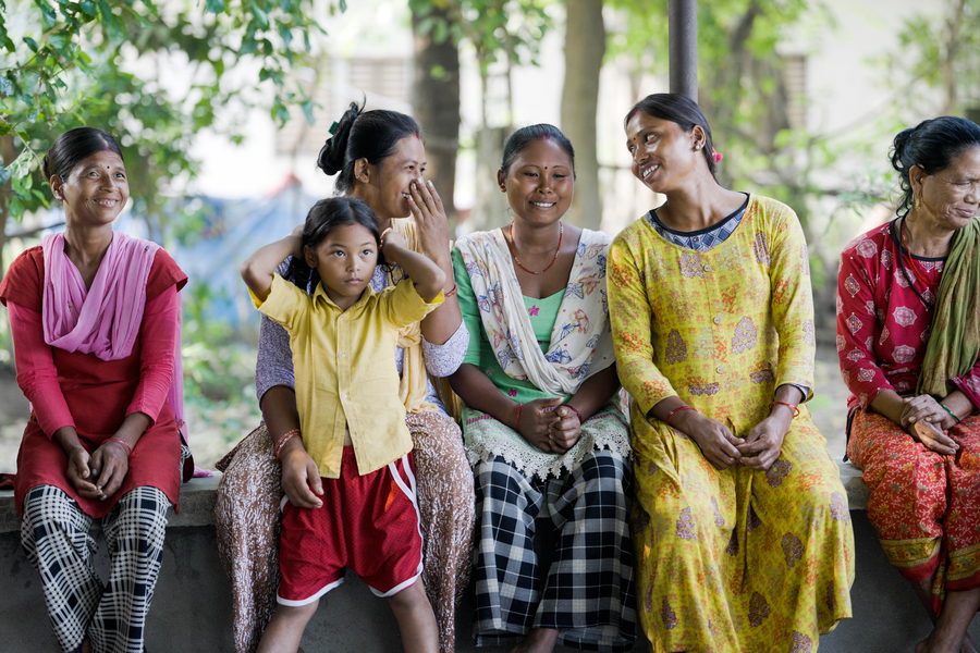A group of women and a child sit together outdoors, sharing smiles and laughter in a vibrant, communal setting.
