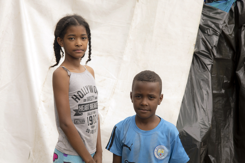 A girl in a grey tank top with writing on it and a boy in a blue football jersey standing next to a tent look to camera.