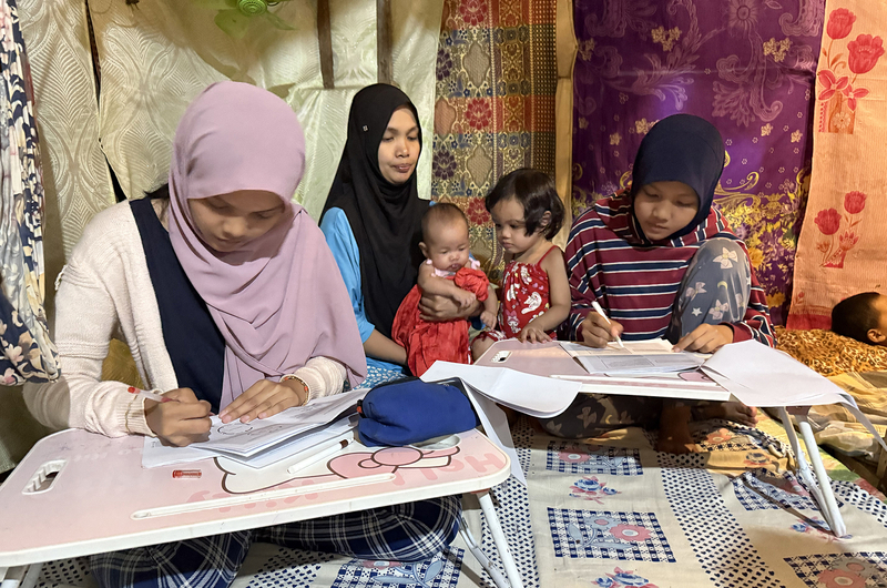 Several family members sit together inside a home while completing paperwork at small study tables.