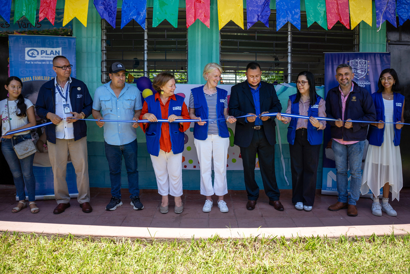 A group of people stand in a line holding a ribbon across the front of a building. Several of them are wearing blue vests with the Plan International logo.