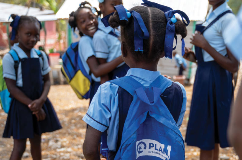 A group of girls in Haiti wearing school uniforms; one girl with her back to the camera wears a Plan-branded backpack filled with school supplies.