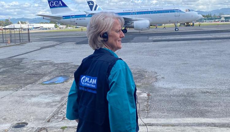 A woman wearing a blue dress is standing on an airport tarmac with a plane in the background.