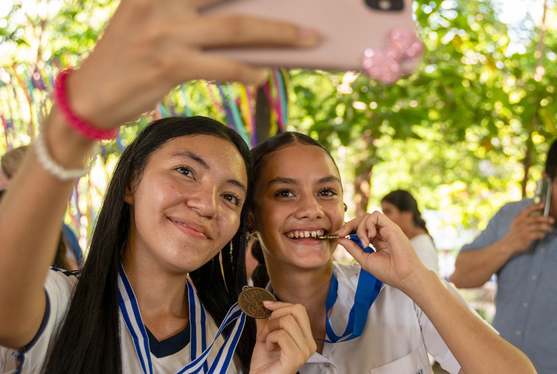 Two schoolgirls smile and take a selfie, wearing medals after a robotics competition.