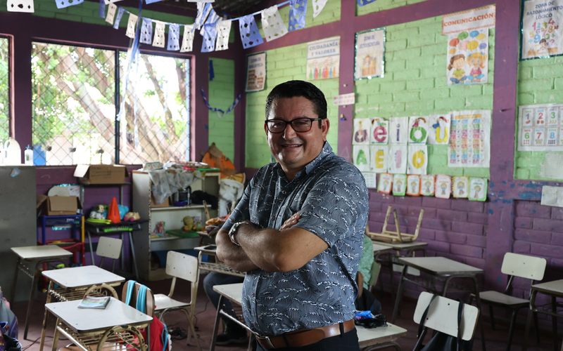  A man stands with his arms crossed in the centre of a brightly lit classroom, smiling. Small desks and chairs are arranged in rows, and the walls display educational posters like an alphabet chart and number cards.