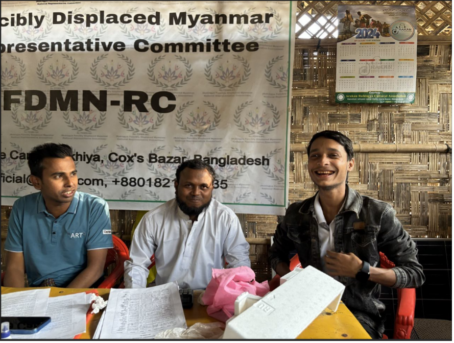 Three men talking and smiling with each other while they’re sitting at a desk.