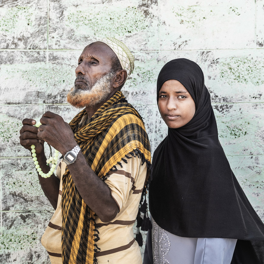 An older man with a beard wearing mostly yellow and brown stands next to a young woman wearing a black headscarf.