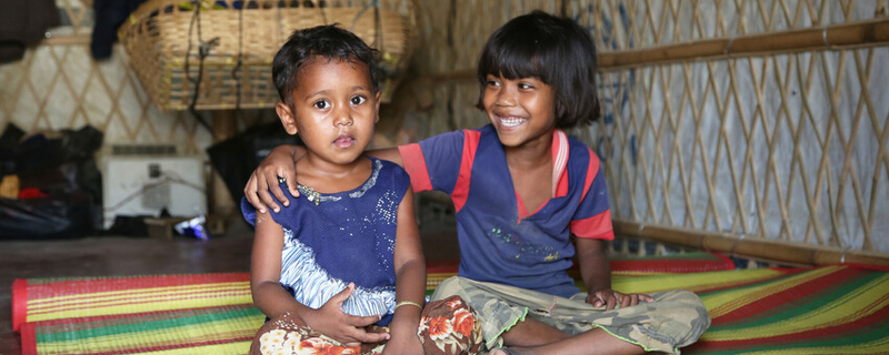 Two little girls sit on a mat with their arms around each other.