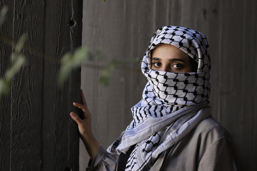 A woman, the lower part of her face covered by a keffiyeh, looks at the camera. 