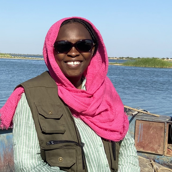 A woman wearing a casual outfit and a pink headscarf sitting on the edge of a boat with a body of water behind her.