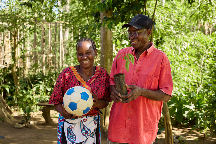 A woman stands smiling while holding a soccer ball beside a man, who is also smiling, holding a seedling in a paper bag.