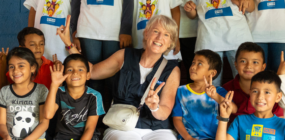 A group of about 20 smiling children in Guatemala holding their hands up and making peace signs, posing with Plan International Canada CEO Lindsay Glassco.